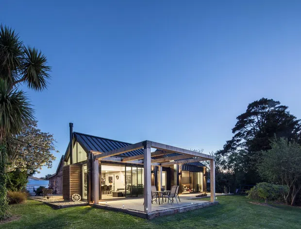 Exterior view of a compact contemporary house or pavilion set within a landscaped garden. The building has a steep dark metal gable roof, timber-clad walls, extensive floor-to-ceiling glazing, and a covered outdoor living area framed by a dark timber or metal pergola. Mature trees and lawn surround the structure.