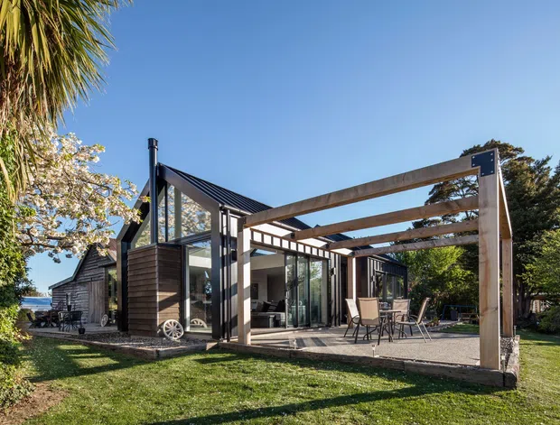 Exterior view of a contemporary single-storey house set in a landscaped garden. The dark-clad home has steep pitched roof forms, extensive glazing, a timber pergola over a paved outdoor dining area, and a partially visible outdoor lounge or living space opening onto the garden.