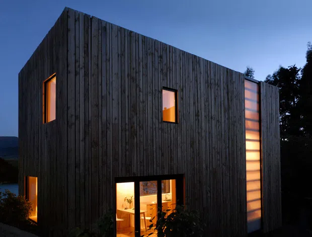 A compact contemporary two-storey house photographed at dusk, with a sharply angular gabled form clad in vertical dark timber boards. Warm light glows through a tall translucent vertical window strip and smaller rectangular windows, while the surrounding landscape and silhouetted trees create a secluded, dramatic setting.