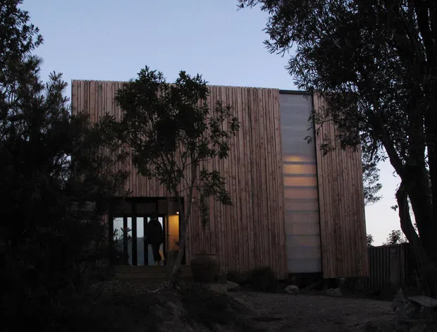 A dark, atmospheric exterior view of a compact modern building set among mature trees. The rectilinear facade is clad in vertical timber boards, with a narrow translucent or glazed vertical section and a glazed entry projecting near the lower left. The surrounding woodland and subdued dusk lighting create a secluded, minimalist character.
