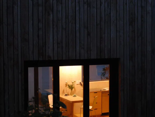 A nighttime exterior view of a compact timber-clad building with dark vertical board cladding and black-framed windows and glazed doors. Warm interior light reveals a small kitchen or dining area and furnishings, while a timber deck and silhouetted planting occupy the foreground.