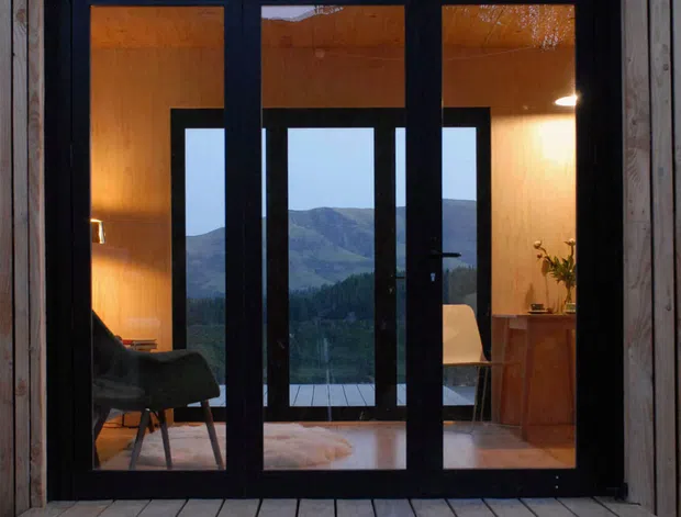 A dark-framed glazed entry or opening set into a vertical timber-clad cabin facade, viewed from a timber deck. Warmly lit interiors are visible through the glazing, including seating, a small cabinet, and a pale rug. Beyond the interior, a forested mountainous landscape and lake or valley are visible. The image has a quiet, rustic contemporary character with strong contrast between black frames, warm wood, and the cool exterior landscape.