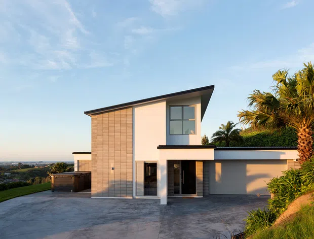 Modern two-storey hillside residence with a sharply sloping roof, pale rendered walls, vertical timber-look cladding, and large glazed openings. The home sits on a sloped landscaped site with a broad concrete driveway and garage, overlooking a coastal or waterfront landscape in the distance.