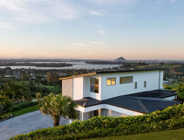 A modern two-storey white-clad home viewed from an elevated landscaped setting, with dark low-pitched roofing, expansive glazing, and a broad paved driveway. The residence overlooks a suburban and coastal or estuarine landscape with water, distant shoreline, and a distinctive volcanic-looking hill beyond. Dense hedging, palms, and manicured planting frame the foreground.