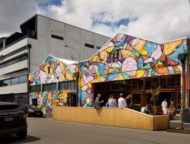 Street-level exterior view of a contemporary commercial building with a vivid multicoloured abstract mural across its angular facade. The open-front venue includes large glazed openings, a timber-clad outdoor dining terrace, and visible patrons, with a larger modern concrete and glazed building adjoining it.