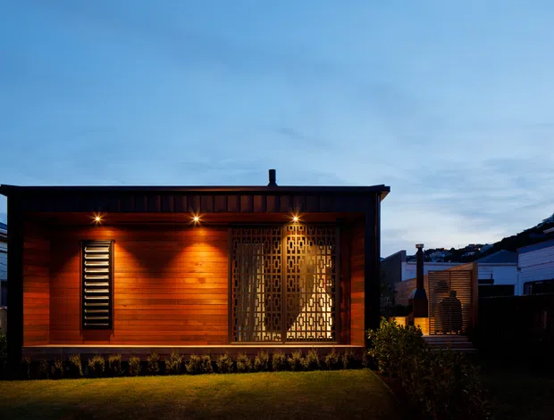 A compact single-storey contemporary dwelling at dusk, clad in warm horizontal timber boards with a dark roof edge and illuminated recessed porch. A narrow louvered window and decorative geometric metal privacy screen flank the entry or living glazing. The house sits on a small lawn with neighbouring residential structures visible on either side.