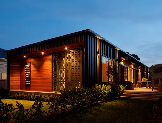 Twilight exterior view of a compact contemporary single-storey dwelling with a dark metal-clad form, warm timber-lined recessed entry, decorative geometric privacy screen, illuminated windows, and a small landscaped garden with a timber deck and outdoor entertaining area.