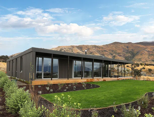 A single-storey contemporary house set in a dry, mountainous landscape. The dark-clad rectangular building has a low-pitched roof, extensive floor-to-ceiling glazing along the front, a covered veranda or terrace, and timber decking. Carefully landscaped garden beds and a broad lawn surround the home, with rugged hills visible behind it.