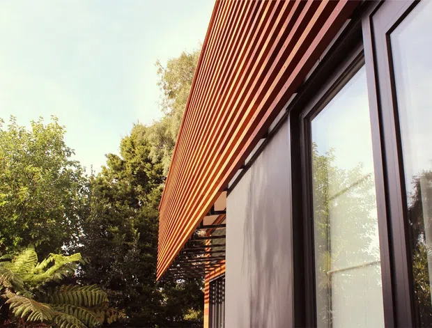 Exterior architectural detail viewed from below, showing a contemporary facade with dark-framed floor-to-ceiling glazing, a projecting upper section clad in closely spaced warm timber battens, and a shaded timber pergola or awning. Dense greenery surrounds the building.