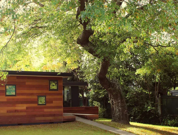 Exterior view of a compact contemporary timber-clad dwelling or extension set among mature trees. The building has horizontal warm-toned wood boards, a low flat roof, dark-framed windows, and a small raised deck or entry platform. A second dark-clad structure is partially visible through the trees.