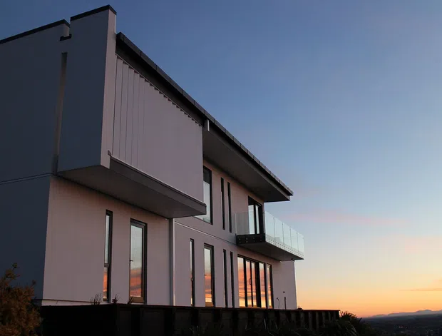 Exterior view of a contemporary multi-storey residence at dusk, with a sharply articulated flat roofline, projecting upper-level volume, smooth pale rendered walls, tall narrow windows, and a glazed balcony. The home overlooks a distant landscape beneath a sunset sky.