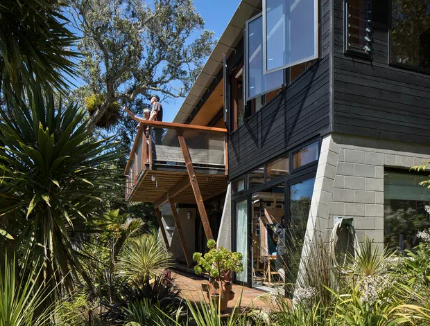 Exterior view of a contemporary multi-level home set within dense landscaped planting. The facade combines dark horizontal cladding, grey masonry block, extensive glazing, and a projecting timber-framed balcony with glass balustrades. A steeply angled roofline and upper-level windows create a strong vertical and geometric composition, while native-style planting surrounds the building.