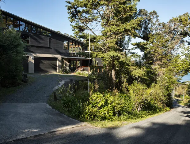 Exterior view of a contemporary multi-level dark-clad house set within dense established vegetation. The building features long horizontal proportions, extensive glazing, a low-pitched roof, upper-level clerestory-like windows, and an integrated garage accessed by a curved driveway. A narrow road runs alongside the landscaped site, with a glimpse of water visible through the trees.