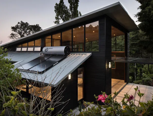Exterior view of a contemporary multi-level house set among dense trees and garden planting. The dark timber-clad structure has a sharply angled roof, extensive floor-to-ceiling glazing, warm illuminated interiors, and a prominent rooftop solar hot-water system. A timber deck and external walkway connect to the raised lower-level entry.