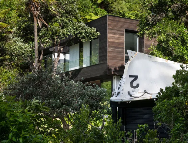 A contemporary elevated residence nestled among dense tropical vegetation. The building features a compact, box-like form with dark horizontal timber cladding, extensive floor-to-ceiling glazing, and a dark roller-style garage or service opening below. Lush palms and mature greenery surround the structure, while a large white boat-like object partially obscures the lower facade.
