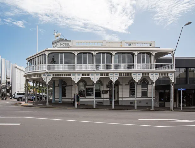 Street-facing photograph of the historic Hart Hotel, a two-storey corner building with an ornate painted veranda, decorative columns, balustrades, arched openings, and classical detailing. A contemporary dark commercial building adjoins it on the right, with a broad urban roadway and streetscape in the foreground.