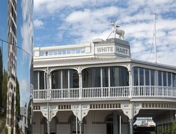 Exterior view of the White Hart building, featuring ornate heritage detailing, a curved glazed façade, decorative balustrades, columns, a rooftop deer sculpture, and a prominent rooftop sign. A modern reflective building is visible at the left edge.