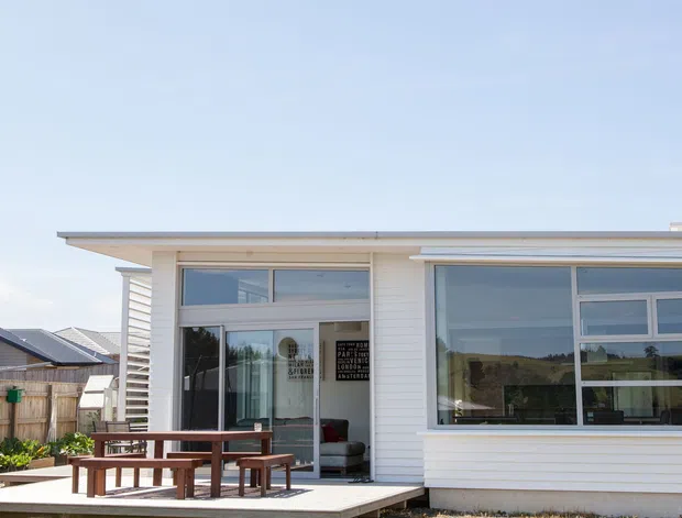 Exterior view of a low-profile contemporary home with white horizontal weatherboard cladding, broad roof overhangs, and extensive aluminium-framed glazing. A raised timber deck with a picnic-style outdoor table occupies the foreground, while neighbouring residential buildings and fencing are visible in the background.
