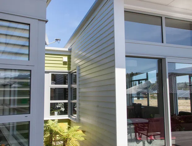 Exterior architectural photograph of a contemporary residential courtyard connection. White horizontal weatherboard-clad volumes frame a glazed living area with large sliding doors, opening onto a timber deck. A narrow planted rock garden with ferns sits between the building wings, and the composition shows clean roof overhangs, white frames, and a light, airy material palette.