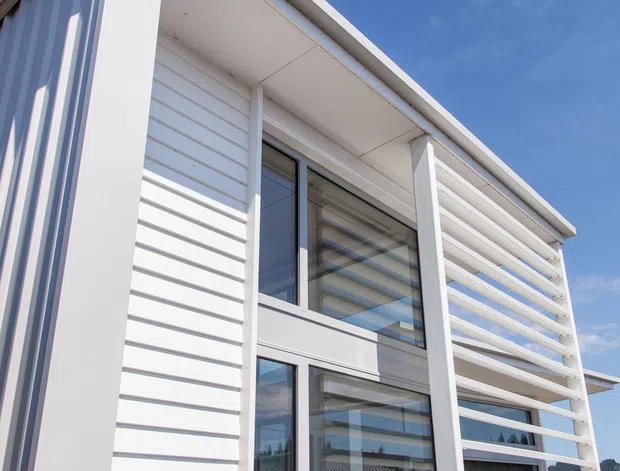 Low-angle exterior view of a contemporary white-clad multi-storey building with horizontal weatherboard-style siding, large dark-framed windows, projecting roof eaves, and a prominent horizontal louvred screen or balcony balustrade. Clear blue sky and a glimpse of outdoor seating are visible.