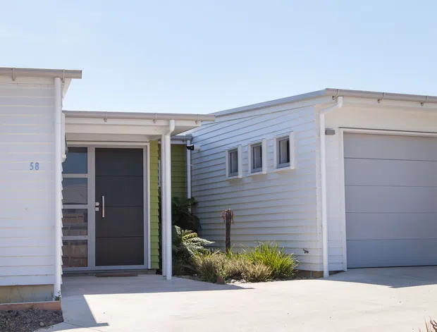 Bright exterior view of a contemporary single-storey residence with pale horizontal weatherboard cladding, a recessed dark entry door, attached single garage, concrete driveway, and low ornamental landscaping. The flat-to-low-pitched roof forms and clean lines create a modern coastal/suburban appearance.