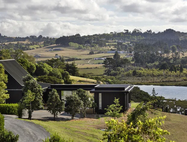 A dark-clad rural property viewed from an elevated position, with a pitched-roof main structure and low flat-roofed connected pavilion in the foreground. The landscaped grounds slope toward a calm waterway, with expansive rolling farmland, trees, and scattered rural buildings beyond under a cloudy sky.