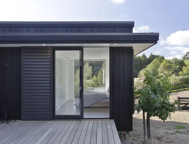 Exterior view of a compact contemporary dark-clad building with a low flat roof and broad covered eaves. A timber deck leads to full-height glazed doors, through which a simply furnished bedroom is visible. The property sits in a leafy semi-rural setting with fencing and trees beyond.
