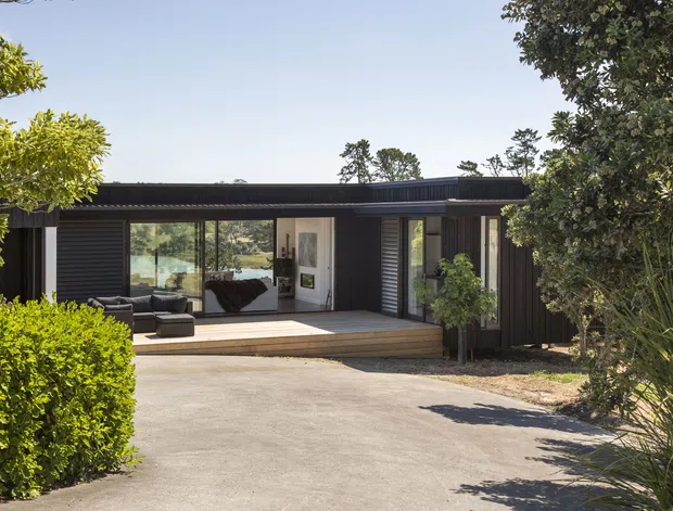 Single-storey contemporary dark-clad residence viewed from the driveway, with a low flat roof, timber deck, broad glazing, and a recessed entry. The home is integrated into a leafy landscaped suburban or rural setting, with a paved approach, hedge planting, mature trees, and ornamental grasses. A living room with dark seating is visible through the glazing.