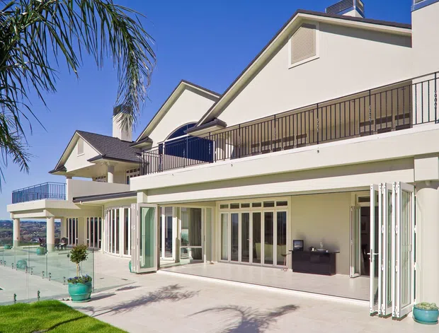 Exterior view of a large two-storey white-rendered residence with multiple gabled roof sections, broad upper-level balconies with dark metal balustrades, extensive folding glazed doors opening onto a paved terrace, and a swimming pool visible at the side. Palm landscaping and bright clear conditions reinforce a resort-like outdoor setting.