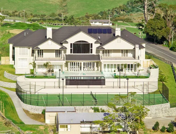 A large symmetrical multi-storey residential estate viewed from above, set on a landscaped sloping site. The house has a dark pitched roof with multiple gabled projections, pale rendered walls, extensive verandahs and glazed openings, a central feature window, and a formal front garden. A fenced tennis court occupies the foreground, with a driveway and rural green surroundings beyond.