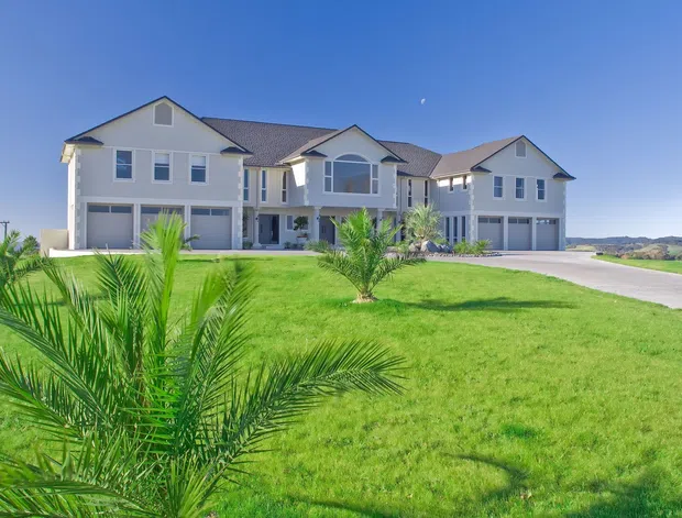 A large, light-coloured multi-section residential building viewed from the landscaped front lawn. The facade features multiple gabled roof sections, white or pale cladding, symmetrical windows, and several attached garage bays. Palm trees and expansive green grass surround the property, with a rural or suburban landscape visible in the distance under a clear blue sky.