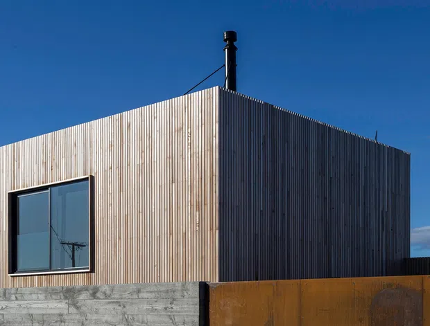 Exterior view of a compact, box-shaped modern building clad in vertically arranged timber battens. A large dark-framed window is set into the front facade, while a black flue projects through the flat roof. The building sits behind a raw concrete wall and a rust-toned metal fence, against a clear blue sky.