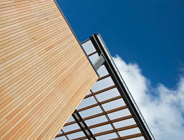 Upward-looking architectural detail showing a modern building facade clad in narrow horizontal timber boards and a projecting pergola or canopy with a dark metal frame and translucent roof panels. Bright blue sky and clouds surround the structure, emphasizing the geometric grid and cantilevered form.