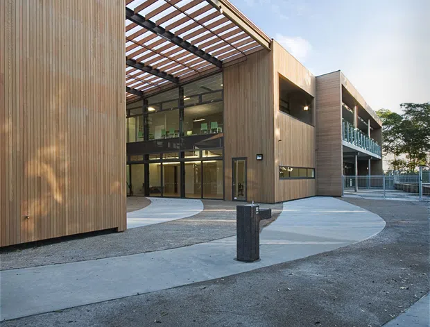 Exterior view of a contemporary multi-storey institutional or commercial building arranged around a paved entry forecourt. The facade combines vertical timber cladding, extensive glazing, exposed steel framing and a translucent or open pergola-like canopy over the entrance. Curved concrete pathways and gravel landscaping lead toward the glazed lobby, with a balcony and external circulation visible on the right.