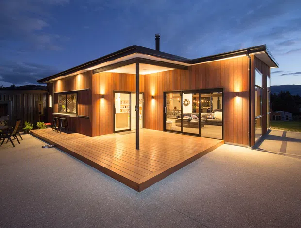 A contemporary single-storey pavilion-style house viewed from an outdoor terrace at dusk. The building has warm vertical timber-clad walls, a low-pitched roof with broad eaves, extensive dark-framed glazing, and a projecting timber deck with illuminated interiors visible through the doors and windows. A paved outdoor area and dining setting surround the main facade.