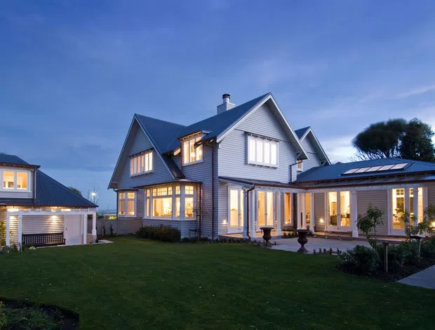 Dusk exterior view of a large, multi-wing home with prominent pitched gable roofs, grey horizontal weatherboard cladding, white trim, and many warmly lit windows. The landscaped lawn and paved approach frame the main residence, with a detached garage or secondary structure visible on the left.