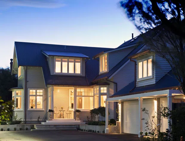 Dusk exterior photograph of a large multi-storey suburban residence with a complex pitched roof, dark corrugated roofing, light horizontal weatherboard cladding, projecting dormer-like window bays, extensive warm-lit glazing, a covered entry porch, front steps, landscaped planting, and an attached double garage or carport structure.