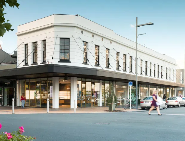 Street-level view of a renovated two-storey commercial corner building with a long white rendered facade, repeated tall industrial-style windows, dark horizontal canopy, and glazed retail frontage at ground level. The building sits on an urban shopping street with pedestrians, parked cars, street furniture, and adjoining shopfronts visible.