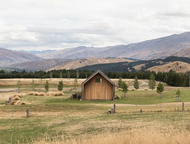 A small timber-clad gable-roofed outbuilding or cabin stands alone in an expansive rural landscape, surrounded by dry grass, pasture, scattered trees, and distant rugged mountains beneath an overcast sky. The composition emphasises the building's simple form and its alpine/rural setting.