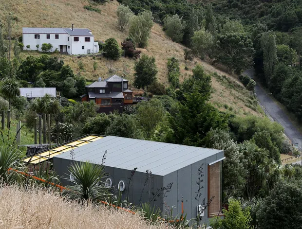 A contemporary low-profile outbuilding with a grey metal roof and dark grey cladding sits on a steep, densely vegetated hillside. Other homes, a narrow winding road, dry grasses, and established trees are visible across the sloping rural landscape. The image is a vertically framed exterior architectural photograph with a muted natural palette.