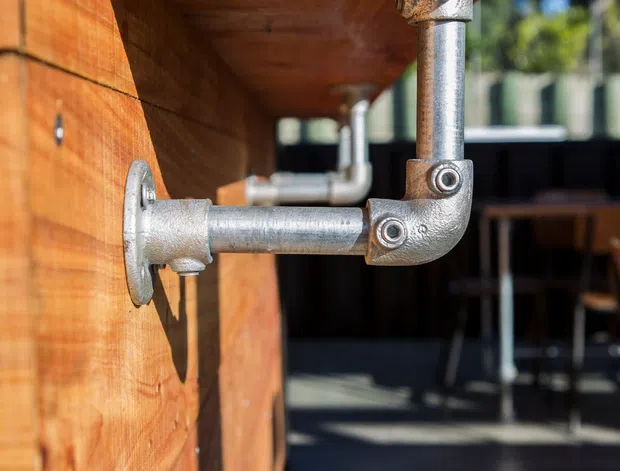 Close-up architectural detail of a thick timber wall and projecting timber shelf or awning supported by exposed galvanized metal pipe brackets and fittings. The image is photographed outdoors in bright sunlight, with a blurred patio or commercial outdoor seating area visible behind it.