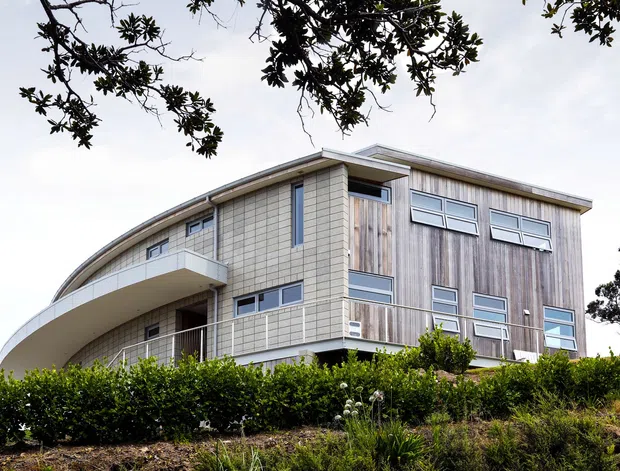 Exterior view of a contemporary multi-storey house on a landscaped, slightly elevated site. The building combines a curved projecting balcony and roofline with pale tiled wall sections, vertical timber-look cladding, extensive rectangular glazing, and slim metal balustrades. Hedging and planted garden beds partially screen the lower level.