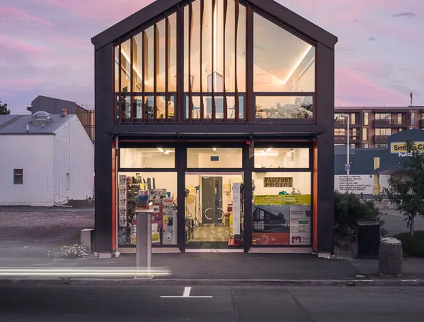 Street-facing urban commercial building photographed at dusk. The compact two-storey structure has a dark gabled frame, extensive floor-to-ceiling glazing on the upper level, and illuminated retail or office spaces at ground level. The scene includes adjacent low-rise buildings, roadway markings, and vehicle light trails, with a pink and blue evening sky.
