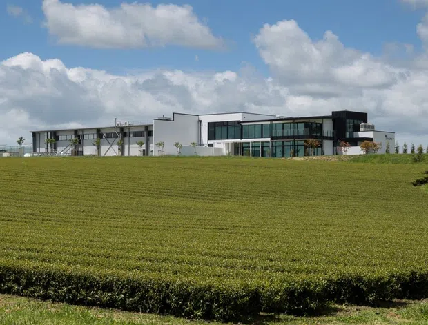 Wide panoramic exterior view of a contemporary low-rise building set behind an expansive neatly cultivated green field. The building has crisp white and dark façades, extensive glazing, a largely flat roofline, and projecting or cantilevered sections. Mature trees and a partly cloudy blue sky surround the rural-looking site.