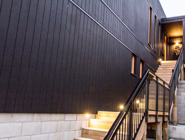 Exterior view of a contemporary multi-level building approached by a long flight of illuminated timber stairs. The facade is clad in dark vertical panels above a light concrete block retaining wall, with black metal balustrades, recessed warm lighting, and a sheltered upper entry. The composition emphasises the building envelope, stair access, and sloped-site response.