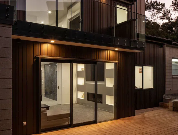 Exterior view of a contemporary multi-level residence at dusk, featuring dark vertical timber cladding, a balcony with black metal framing, large glazed doors, and a warm timber deck. Interior shelving and a bathroom-like space are visible through the ground-floor glazing, with a secondary rendered structure and landscaped steps to the right.