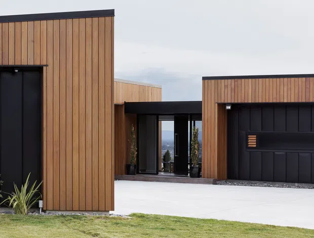 Exterior view of a contemporary multi-volume residence with warm vertical timber cladding, black metal-framed garage and entry doors, and flat rooflines. A covered glazed passage connects the buildings, with a distant coastal or ocean view visible through the entry. Minimal planting and a broad concrete driveway reinforce the clean, modern composition.