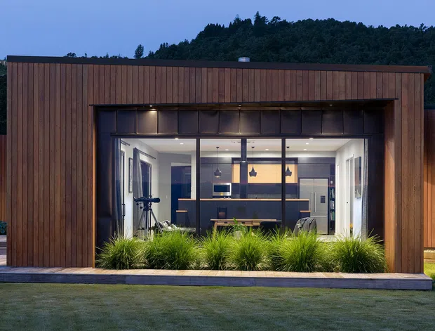 Twilight exterior view of a compact modern pavilion clad in vertical timber, with a large glazed opening revealing a contemporary kitchen and dining area inside. Ornamental grasses line the concrete threshold, and dense wooded hills form the background.