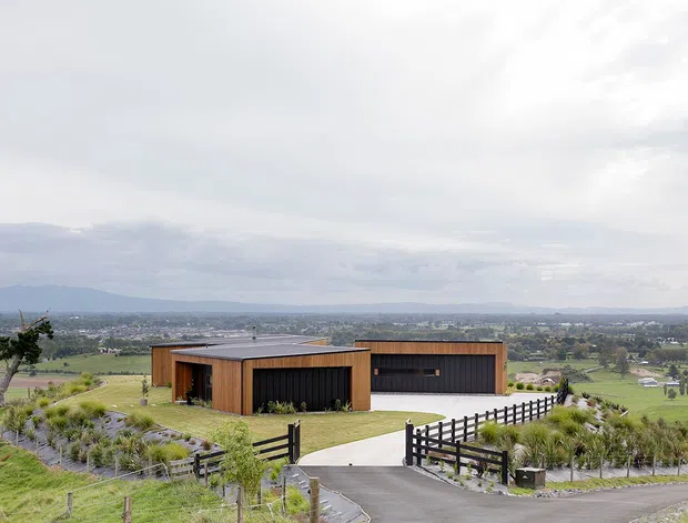 Wide exterior view of a low-profile contemporary house set on an elevated rural property, with long rectilinear volumes, warm timber-clad walls, dark vertical screening, and a flat roof. A driveway and dark timber fencing lead toward the home, overlooking expansive green agricultural fields and distant hills under an overcast sky.