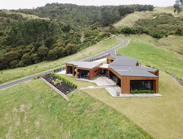 Aerial view of a modern, low-profile residence set within expansive rolling green hills and native woodland. The angular home features dark grey low-pitched roofs, warm timber-clad walls, extensive glazing, paved terraces, landscaped garden beds, and a winding driveway.